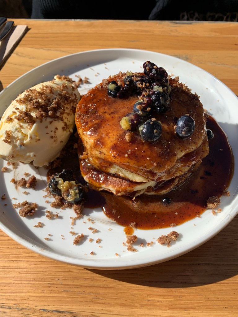 Stack of pancakes cocered in syrup, with blueberries, walnuts and a crumble serves with ice cream